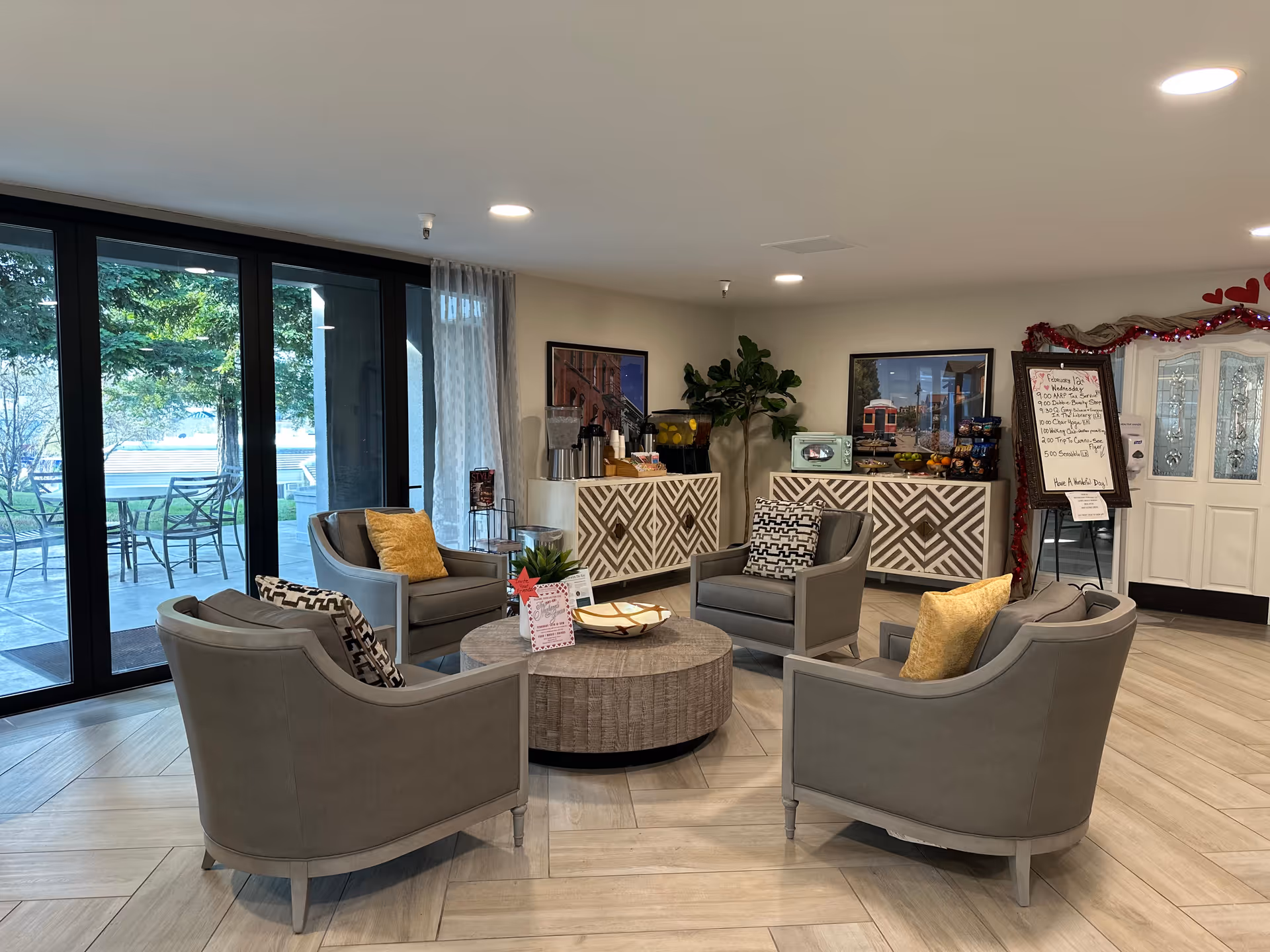 A cozy senior living facility common area with four gray armchairs arranged around a round wooden coffee table. The room has large glass sliding doors leading to an outdoor patio with metal chairs and tables. Inside, there are two patterned cabinets against the wall, a plant, framed pictures, and a whiteboard with a schedule. The floor is light wood, and the space is well-lit with ceiling lights.