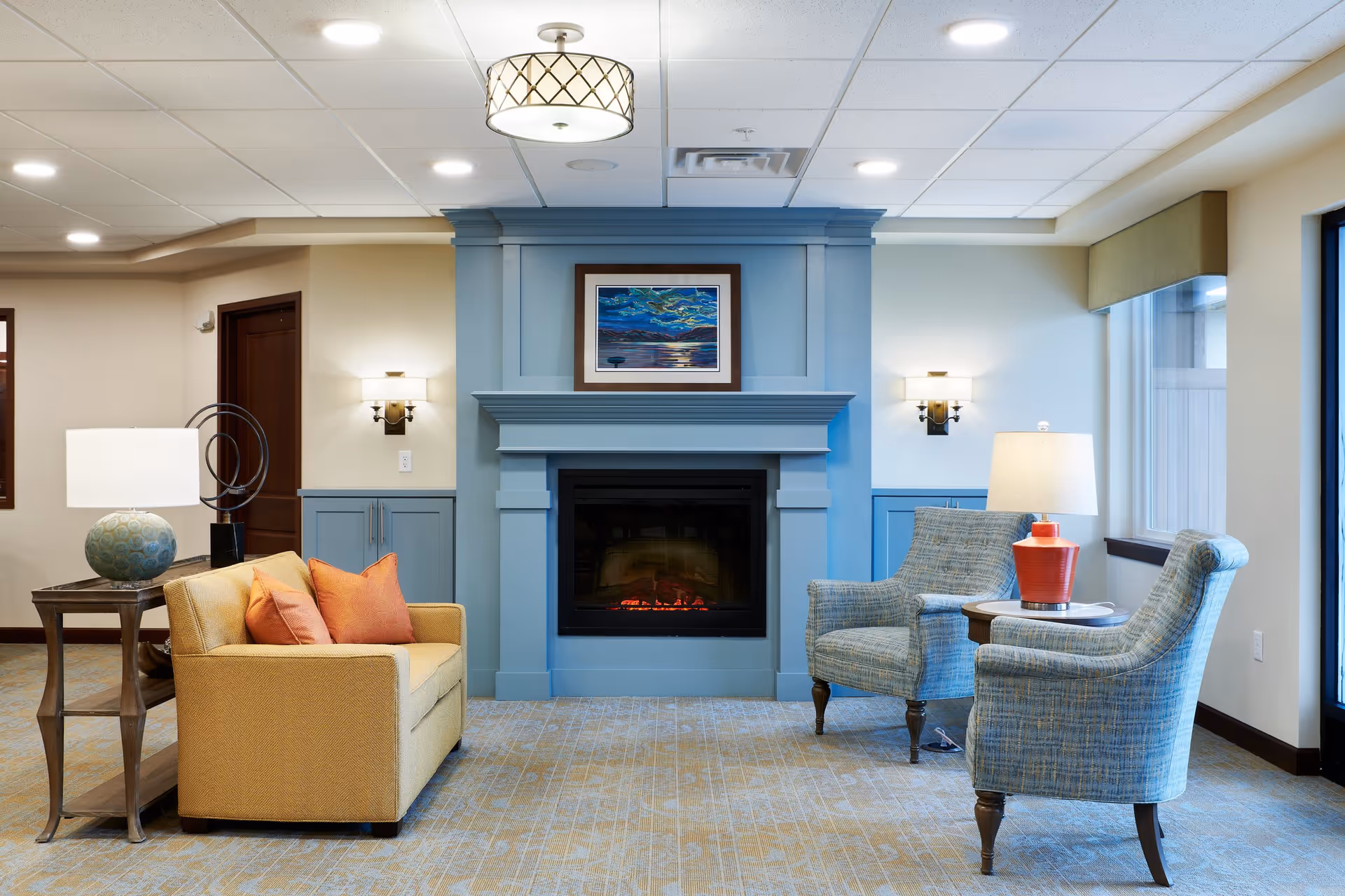 A cozy seating area in a senior living facility with a blue fireplace as the focal point. The fireplace has a framed painting above it. To the left is a yellow loveseat with orange pillows and a side table with a decorative lamp. To the right are two blue upholstered armchairs with a round wooden table and a lamp with a red base between them. The room has soft lighting and a patterned carpet.