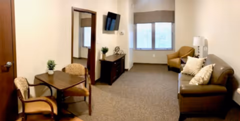 Small furnished living area with a sofa, armchair, wall-mounted TV, side table and a small dining table with chairs.