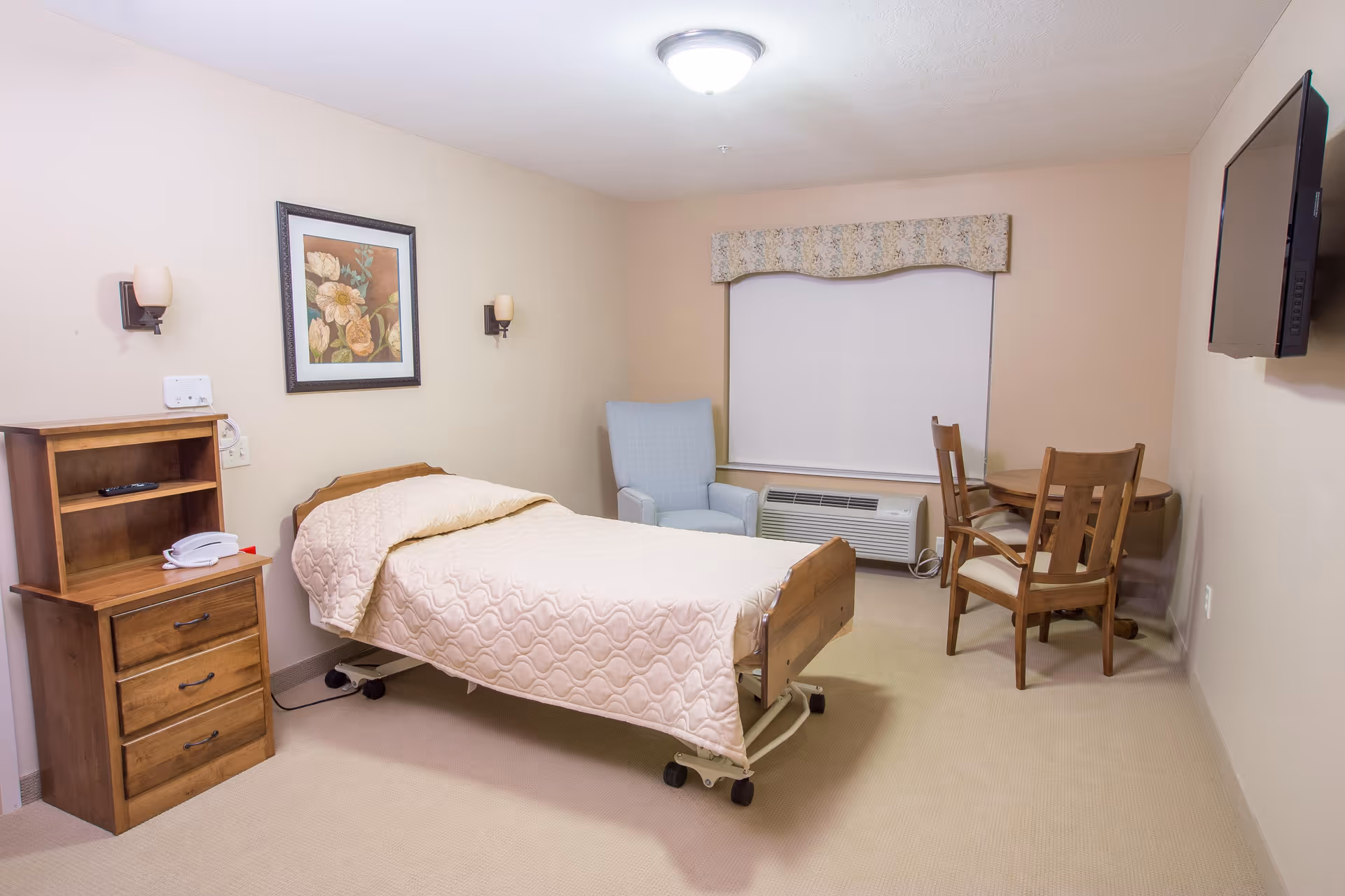 A simple, clean bedroom in a senior living facility with a single bed covered in a beige quilt, a wooden nightstand with a telephone, a light blue armchair, a small round wooden table with two chairs, a wall-mounted flat screen TV, and a window with a floral valance and closed blinds.
