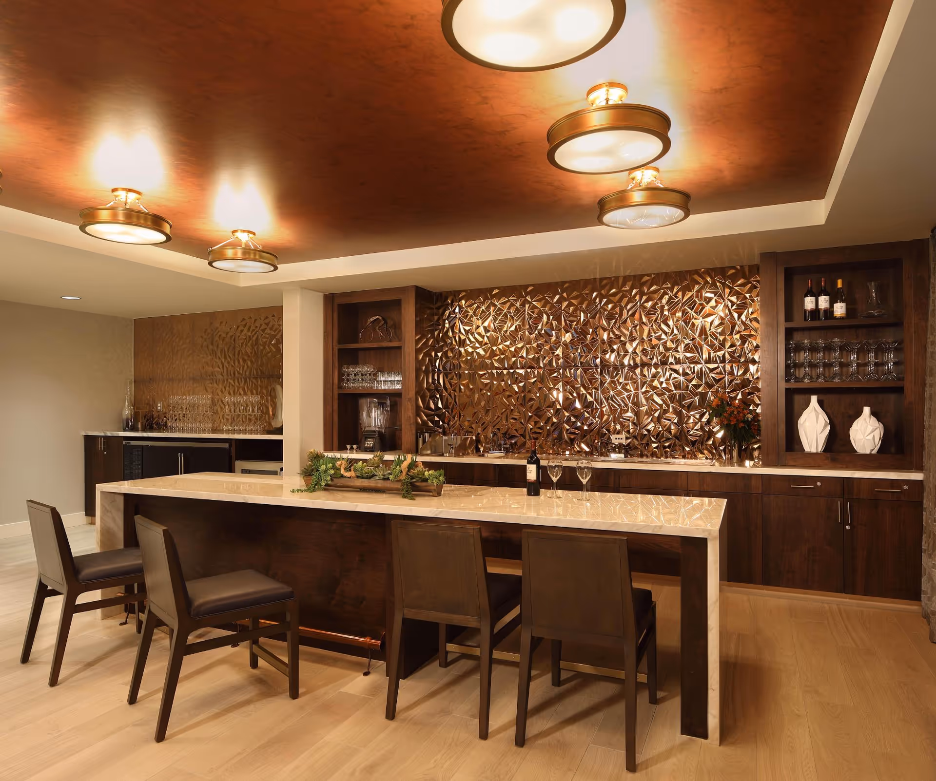Elegant indoor bar/dining area with a marble island, four wooden chairs, decorative bronze geometric backsplash, dark wood cabinetry and pendant ceiling lights.