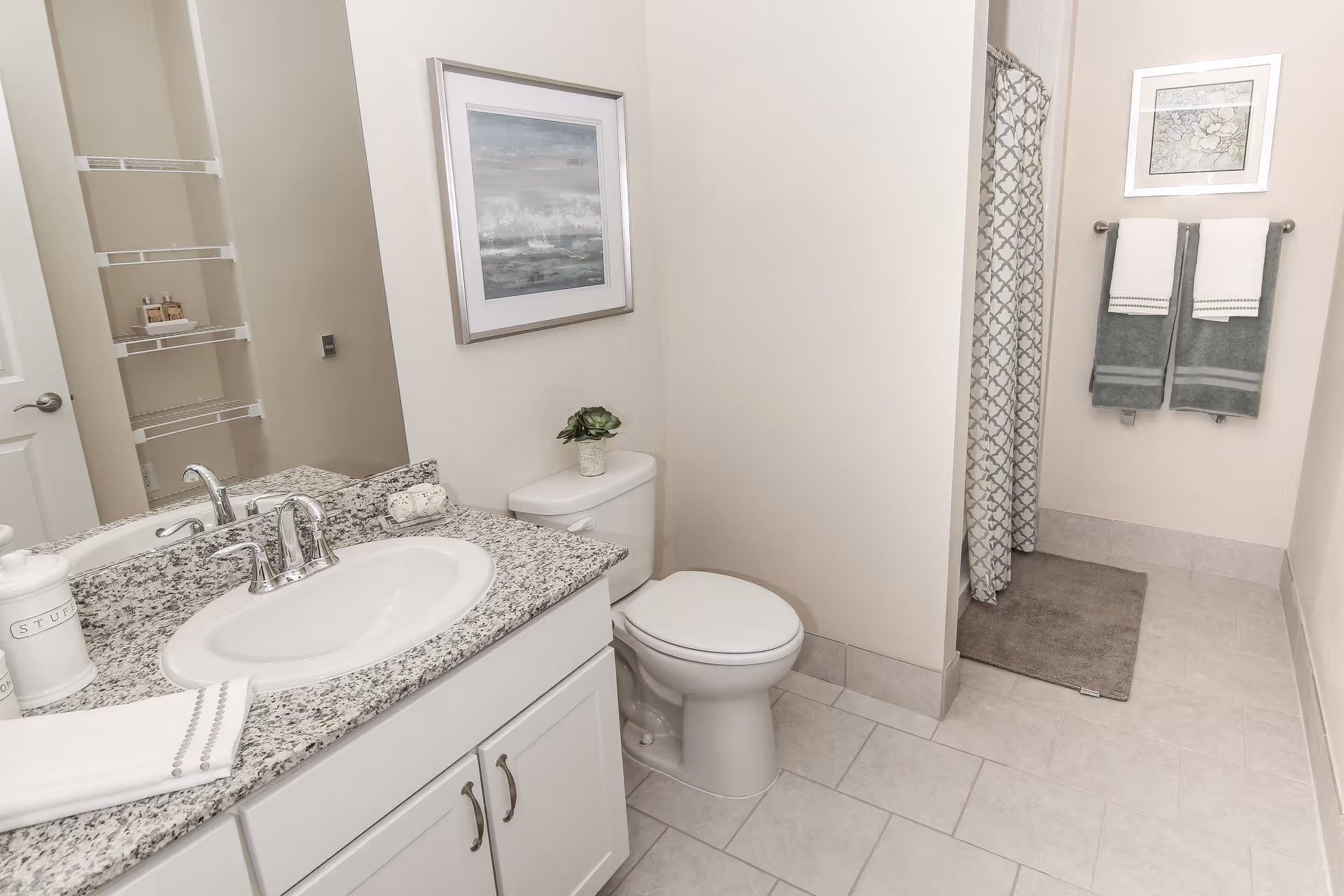 A clean and modern bathroom featuring a white sink with a granite countertop, a toilet with a small potted plant on top, a shower area with a patterned curtain, two green and white towels hanging on a rack, a gray bath mat, and a framed artwork on the wall.