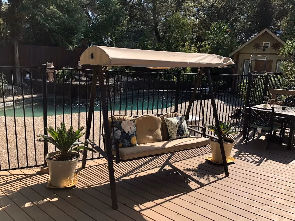Outdoor patio area with a cushioned swing bench with a canopy, two potted plants on either side, a fenced swimming pool in the background, and a small shed or playhouse surrounded by trees and greenery.