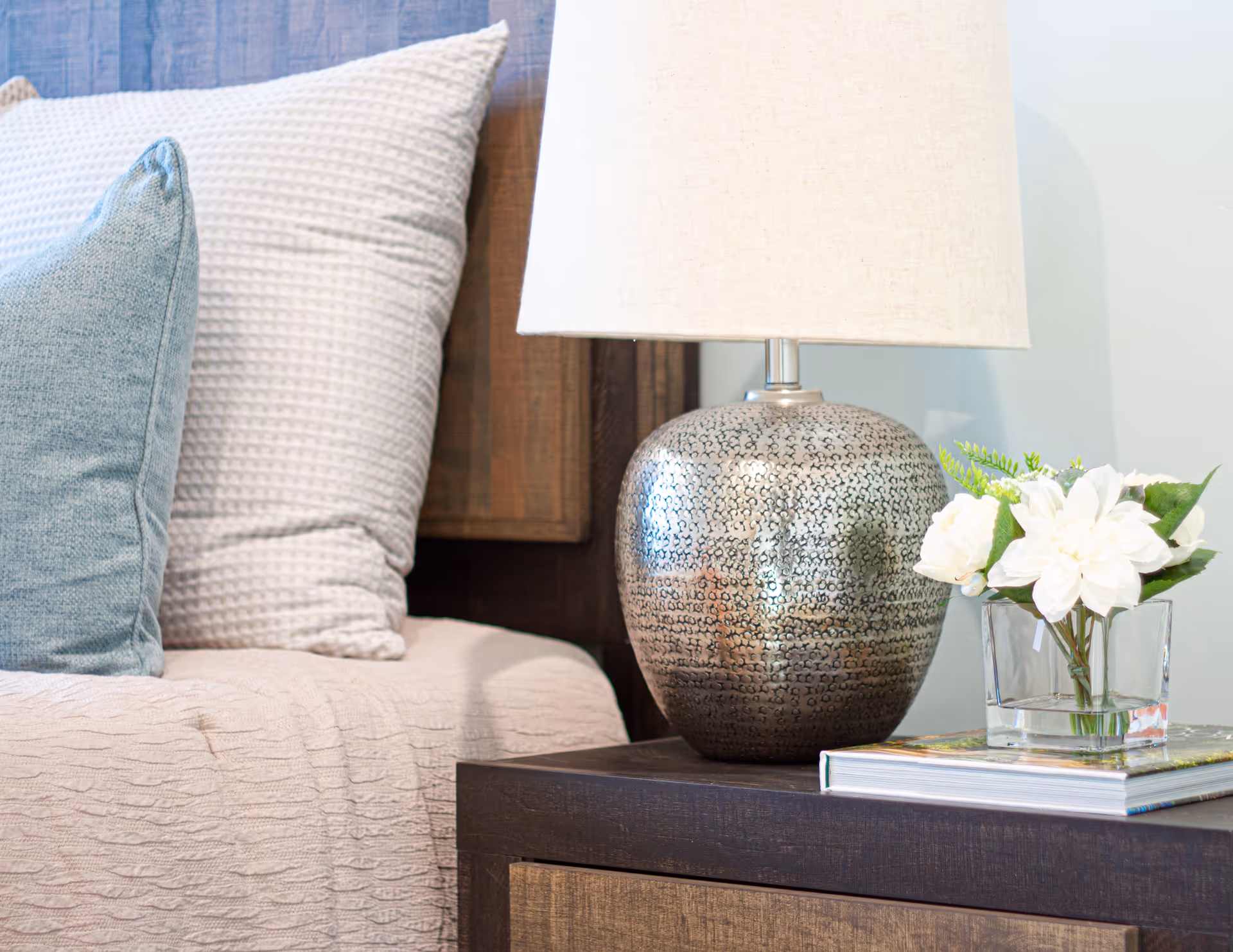 Close-up of a bedside table with a decorative silver lamp, a small glass vase with white flowers, and a stack of books next to a bed with textured pillows and a beige bedspread.