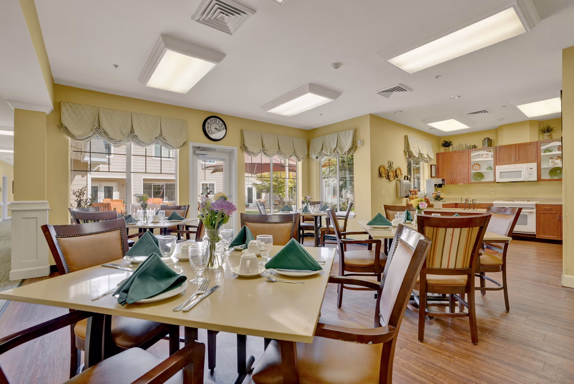 A bright dining room with several tables set with green folded napkins, glassware, and white cups. The room has large windows with light green valances, wooden chairs with cushioned seats, and a kitchen area with wooden cabinets, a microwave, and a dishwasher in the background. The walls are painted yellow and the floor is wood.