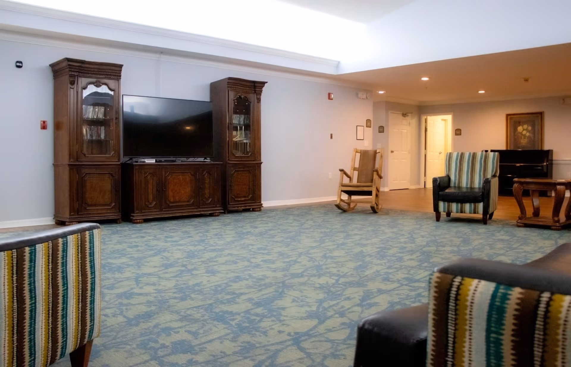 A spacious living room area with a large flat-screen TV set in a wooden entertainment center flanked by two matching wooden cabinets. The room has patterned carpet flooring and several chairs including a wooden rocking chair and striped upholstered armchairs. There is a piano and framed artwork on the far wall, with white doors leading to other rooms.