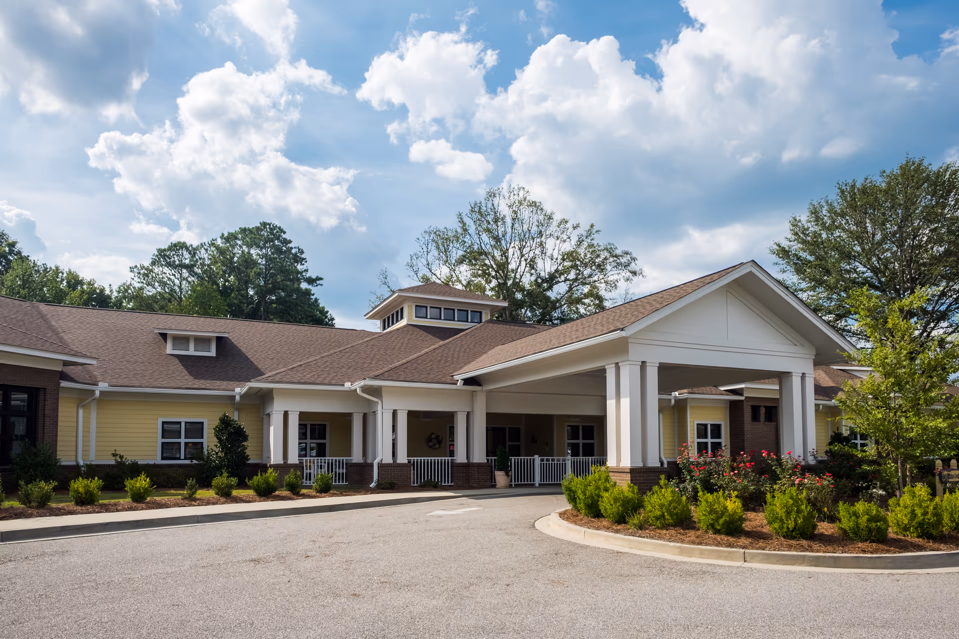 Exterior view of White Oak Manor Rock Hill, showing a single-story building with a covered entrance supported by white columns, surrounded by landscaped bushes and trees under a partly cloudy sky.