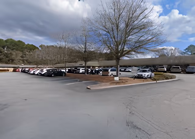 A wide paved parking area with rows of parked cars and golf carts beneath leafless trees under a partly cloudy sky.