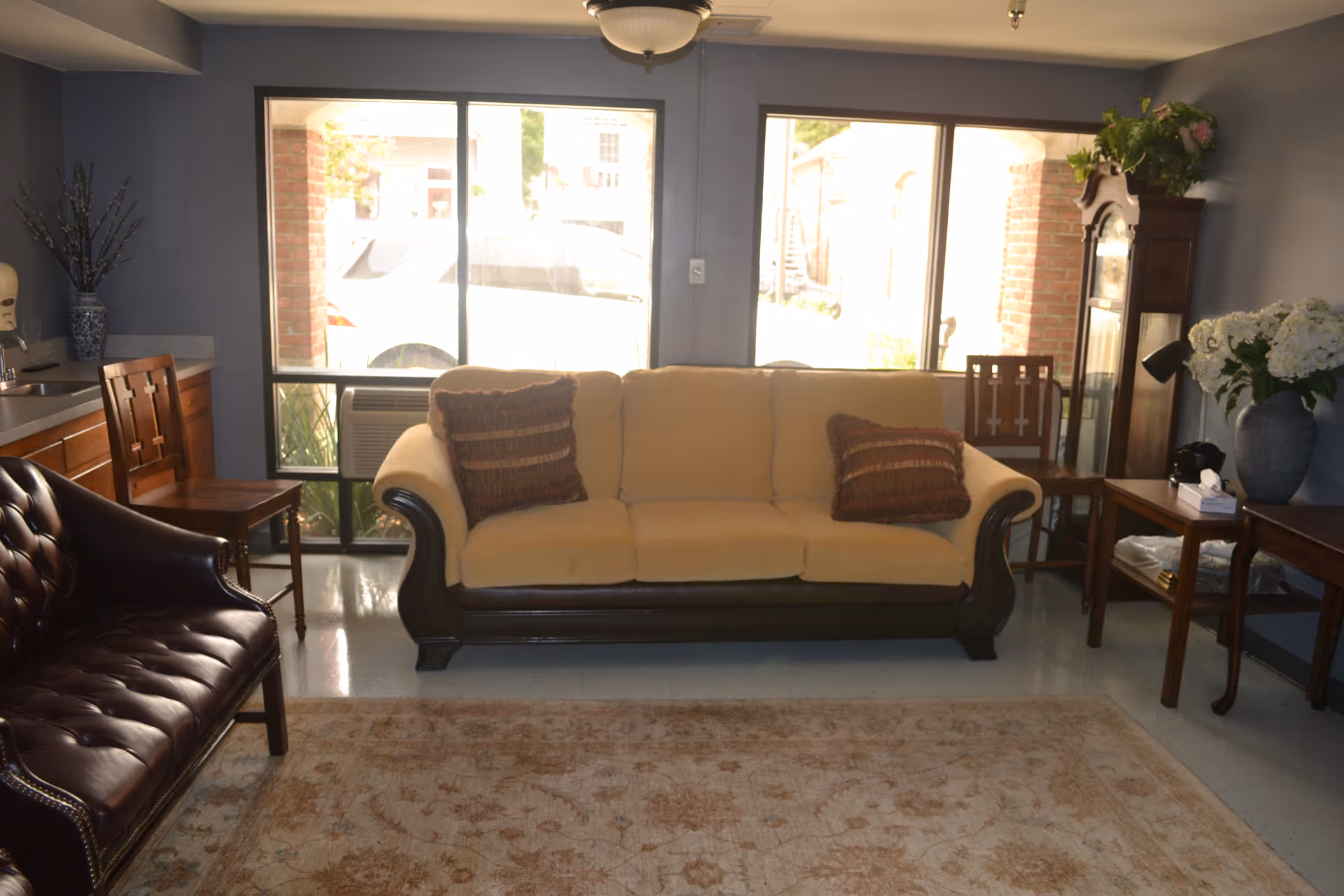 A cozy living room with a beige sofa adorned with two brown patterned cushions, a dark brown leather armchair, wooden chairs, a wooden side table with a vase of white flowers, and a tall grandfather clock. Large windows let in natural light, and a patterned area rug covers the floor.