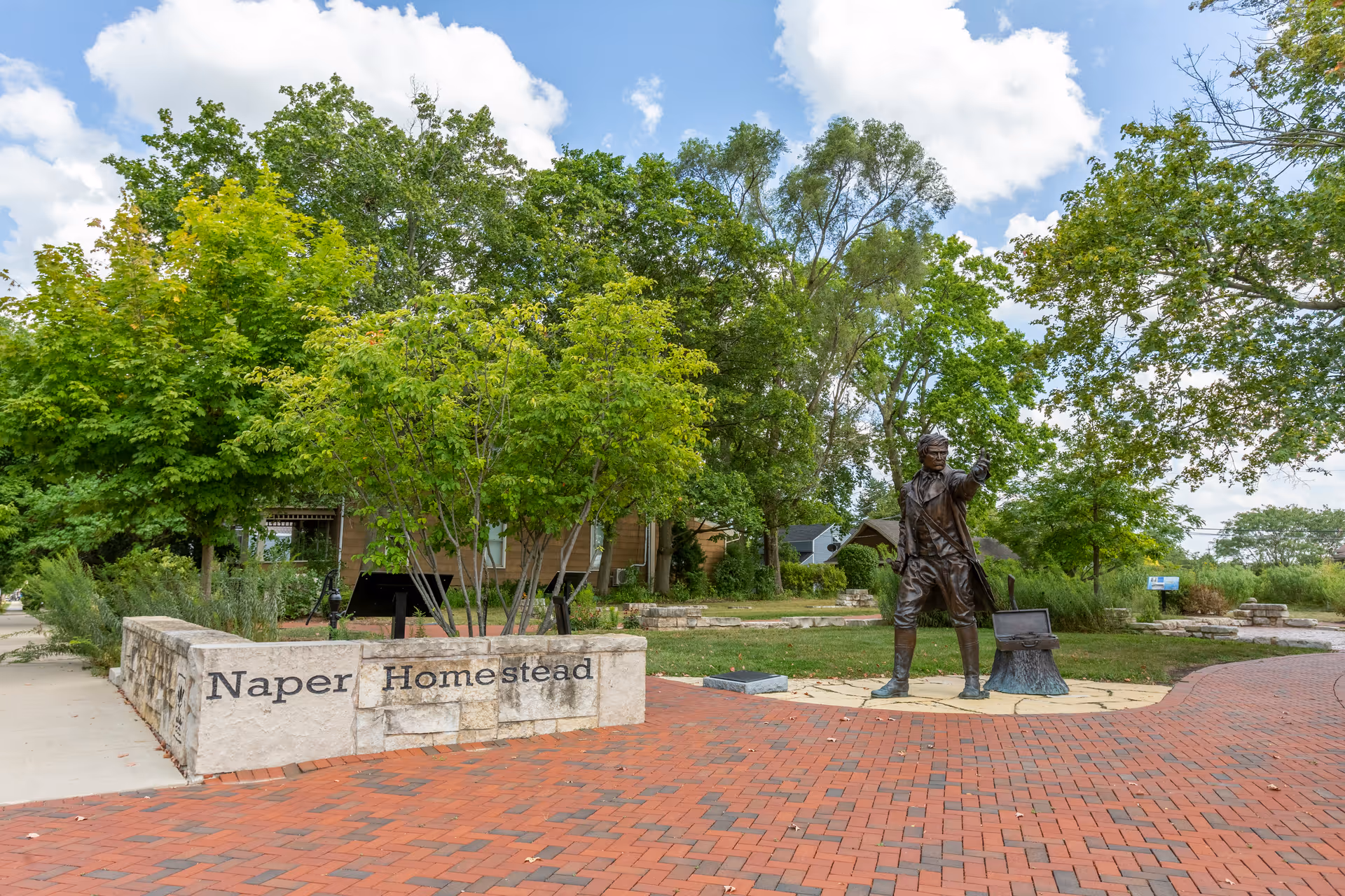 Outdoor scene featuring a brick-paved walkway and a bronze statue of a man pointing forward. The statue stands on a circular stone platform next to a small open chest. Surrounding the area are green trees and shrubs, with a stone wall displaying the text 'Naper Homestead'. The sky is partly cloudy.