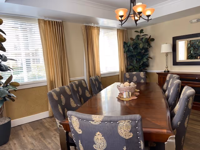 A dining room with a long wooden table surrounded by eight upholstered chairs with patterned fabric. The room has large windows with beige curtains, a chandelier with six lights hanging above the table, a sideboard with a lamp and a mirror on the wall, and two large potted plants in the corners.