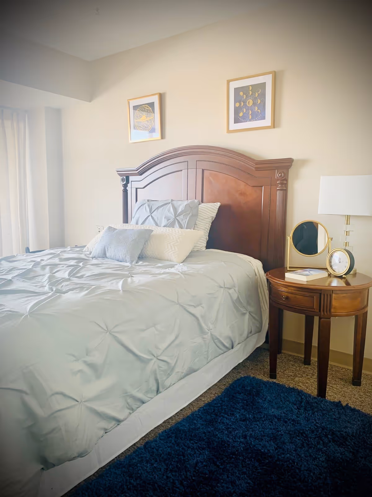 A neatly made bed with a wooden headboard in a bedroom. The bed has light blue bedding and several pillows. Next to the bed is a wooden nightstand with a round mirror, a clock, a book, and a lamp. Two framed pictures hang on the beige wall above the headboard. A blue rug is partially visible on the floor.