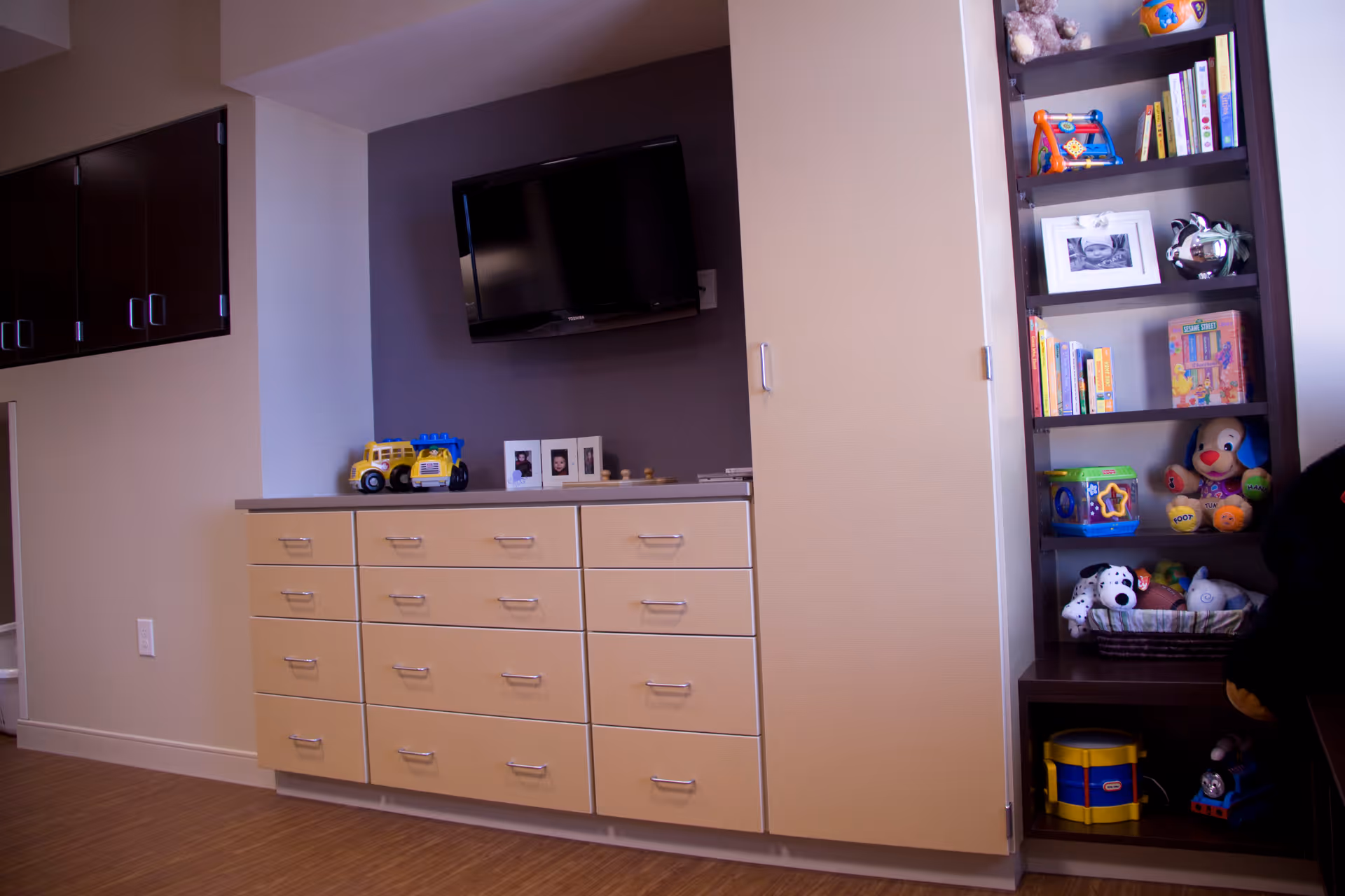 Wall-mounted TV above a bank of drawers and cabinets with shelving filled with toys and books in an interior room.