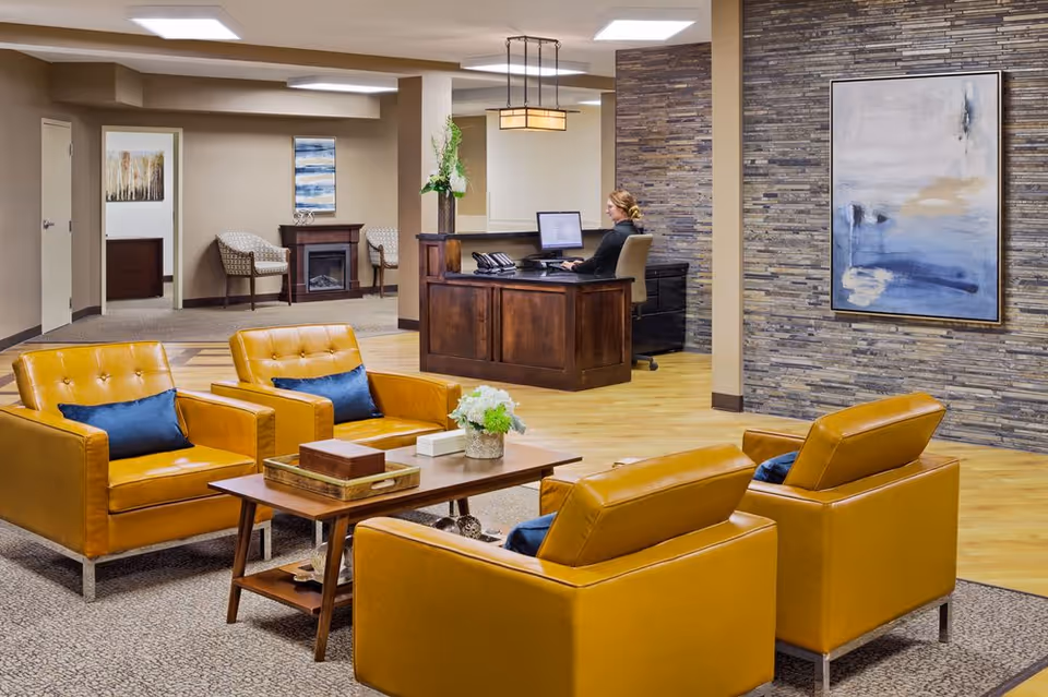 A modern senior living facility lobby with four mustard yellow armchairs arranged around a wooden coffee table with decorative items. A woman is seated at a wooden reception desk working on a computer. The room features a stone accent wall with abstract artwork, light wood flooring, and neutral-colored walls. In the background, there are two patterned chairs and a small electric fireplace.