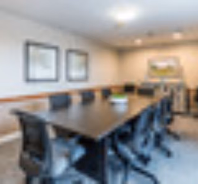 Long conference-style table surrounded by black office chairs in a neutral meeting room with framed artwork on the walls.