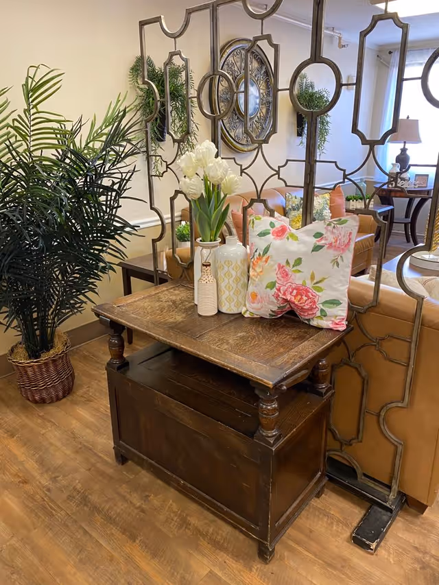 Wooden side table with vases and a floral pillow in a cozy seating area separated by a decorative metal room divider and a potted plant.