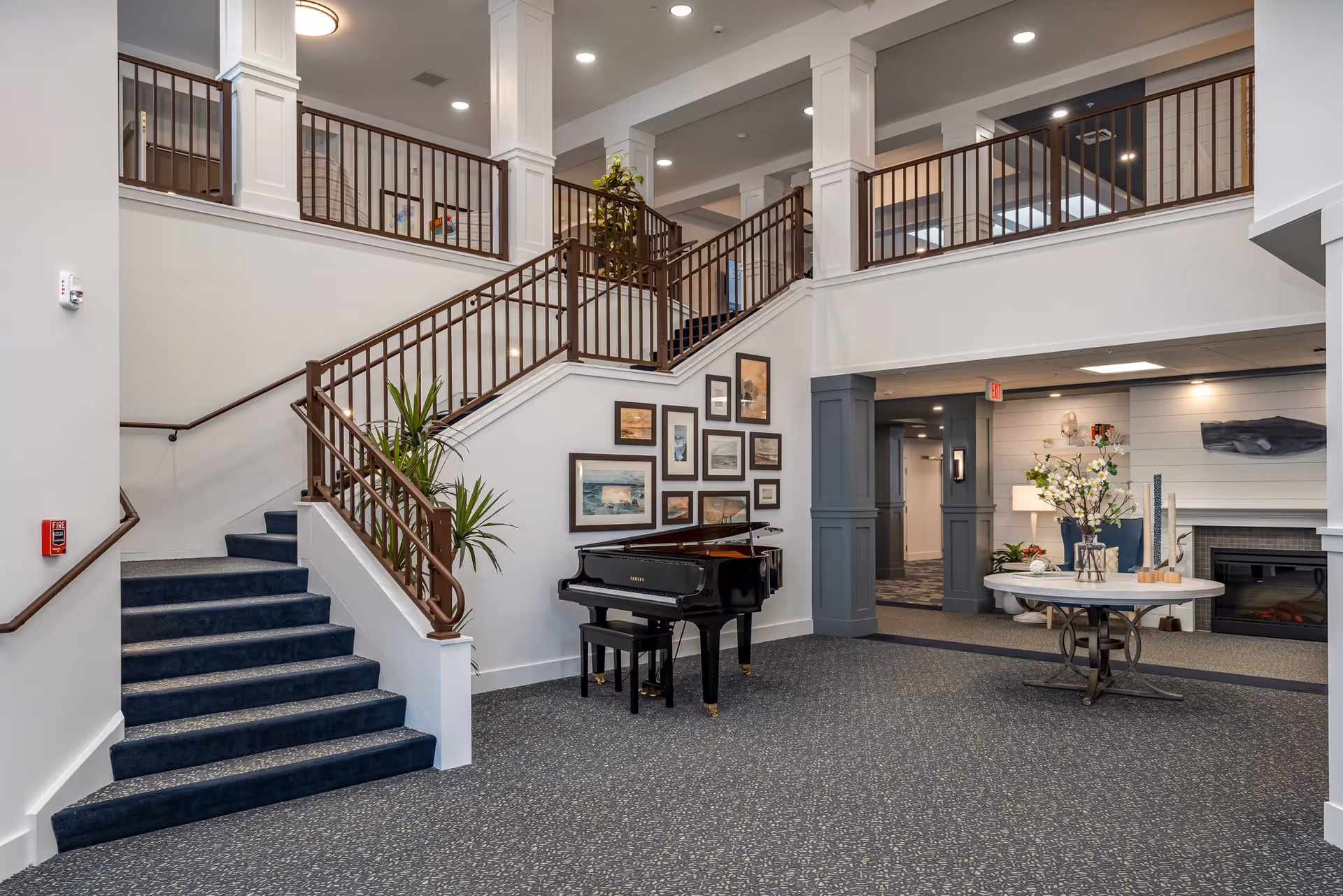 Bright two-story senior living lobby featuring a carpeted staircase, a black grand piano, and a round table with flowers.