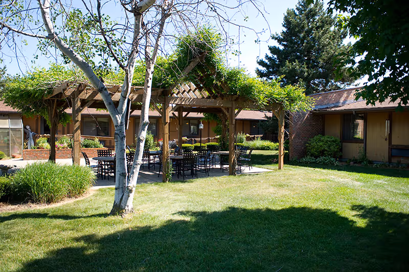 Outdoor garden area at The Village of Westland featuring a wooden pergola with green vines, several black metal tables and chairs underneath, surrounded by grass, trees, and shrubs, with single-story buildings in the background.