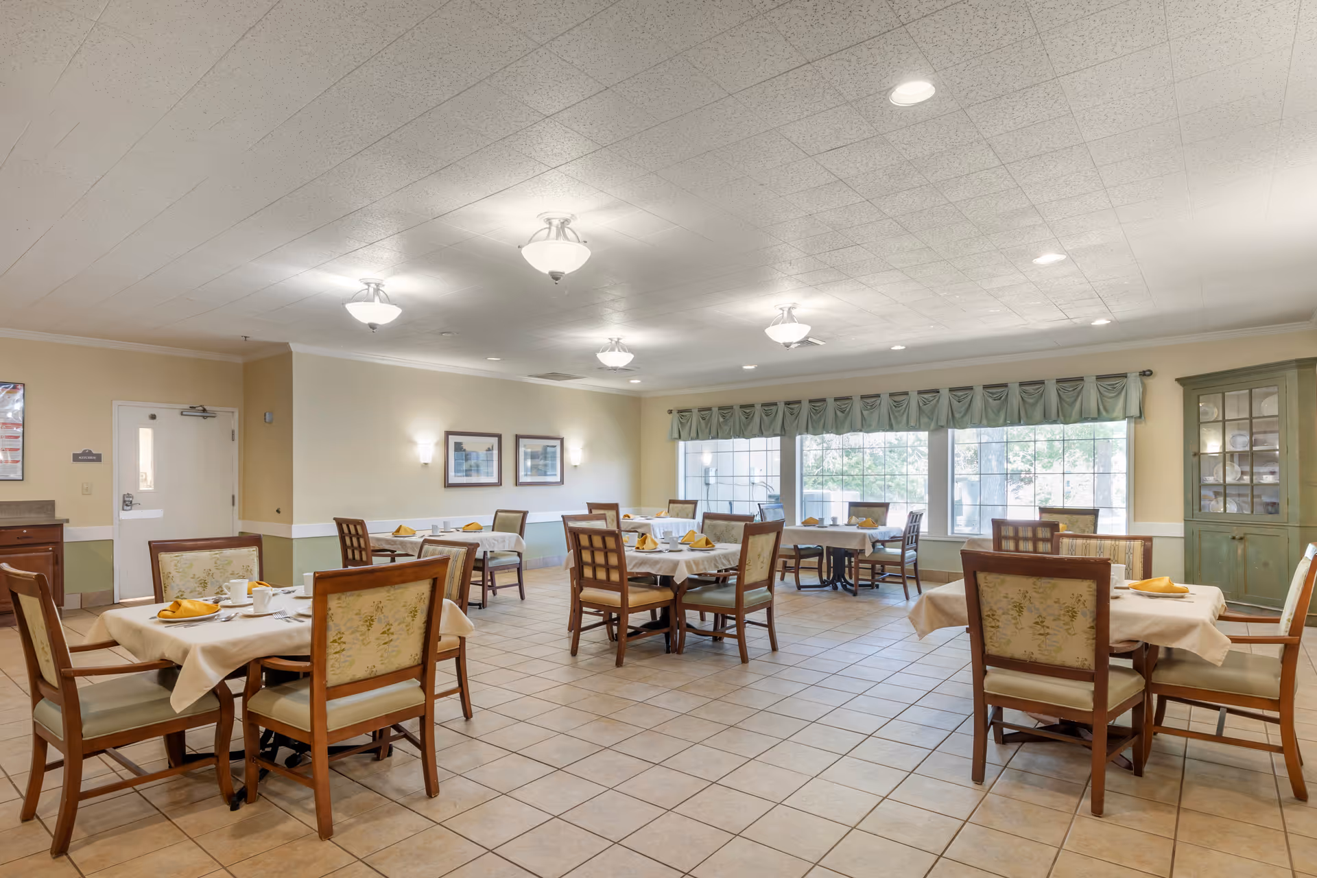 Bright communal dining room with multiple set tables and chairs, large windows, and tiled floor.