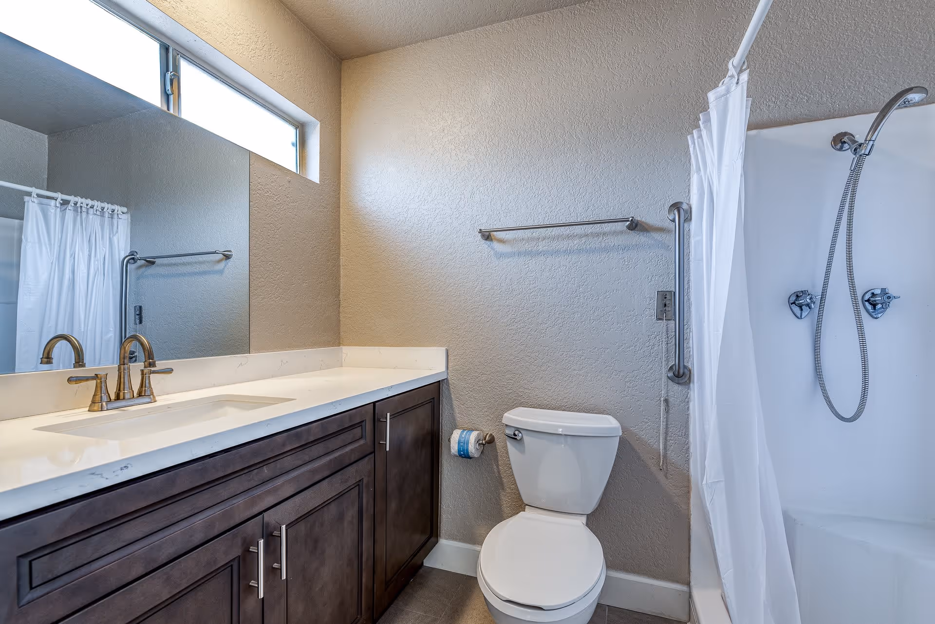 A clean bathroom featuring a white toilet, a dark wood vanity with a white countertop and a sink with brass faucets, a large mirror above the vanity, a towel rack on the wall, and a shower area with a white curtain and a handheld showerhead.