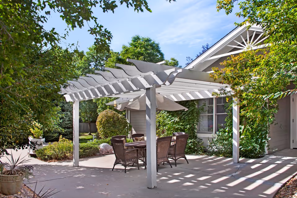 A shaded outdoor patio with a white pergola, wicker dining table and chairs, and lush greenery beside a building.