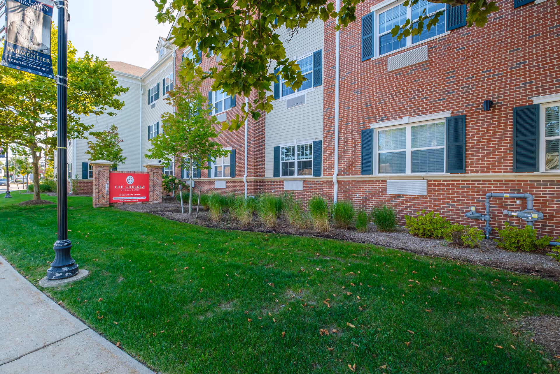 Exterior view of The Chelsea at Fair Lawn building with a red sign in front indicating it is an independent living facility. The building has brick and siding walls with multiple windows and green shutters. There is a well-maintained lawn and some small trees and shrubs along the sidewalk.