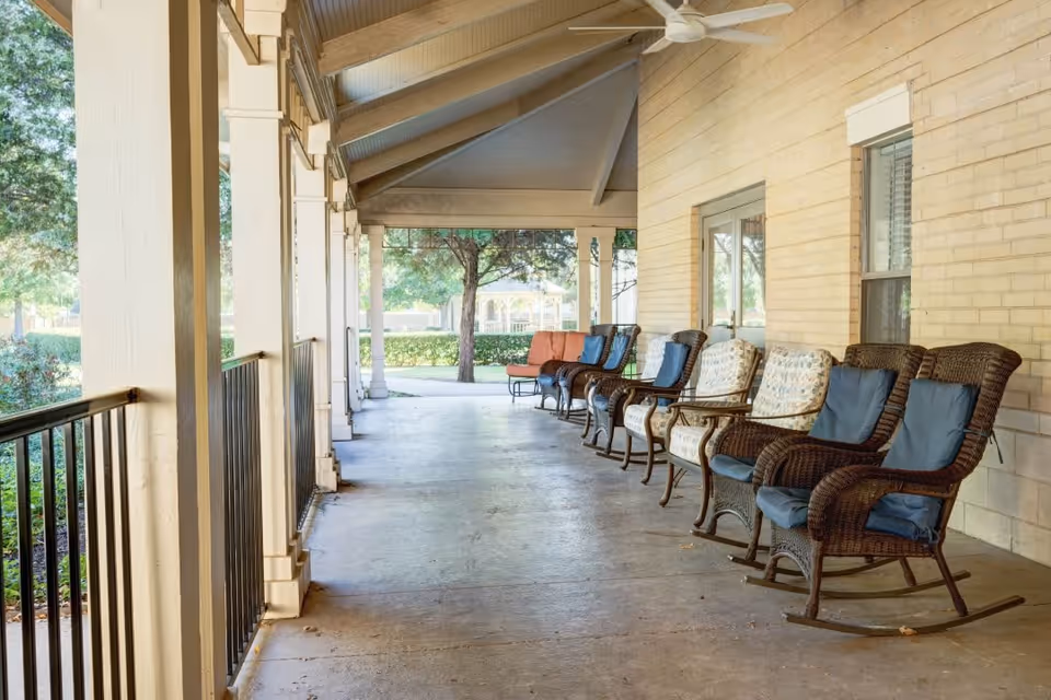 Covered outdoor porch area with a row of wicker rocking chairs and cushioned armchairs lined up against a beige brick wall, overlooking a garden with trees and a gazebo in the background.
