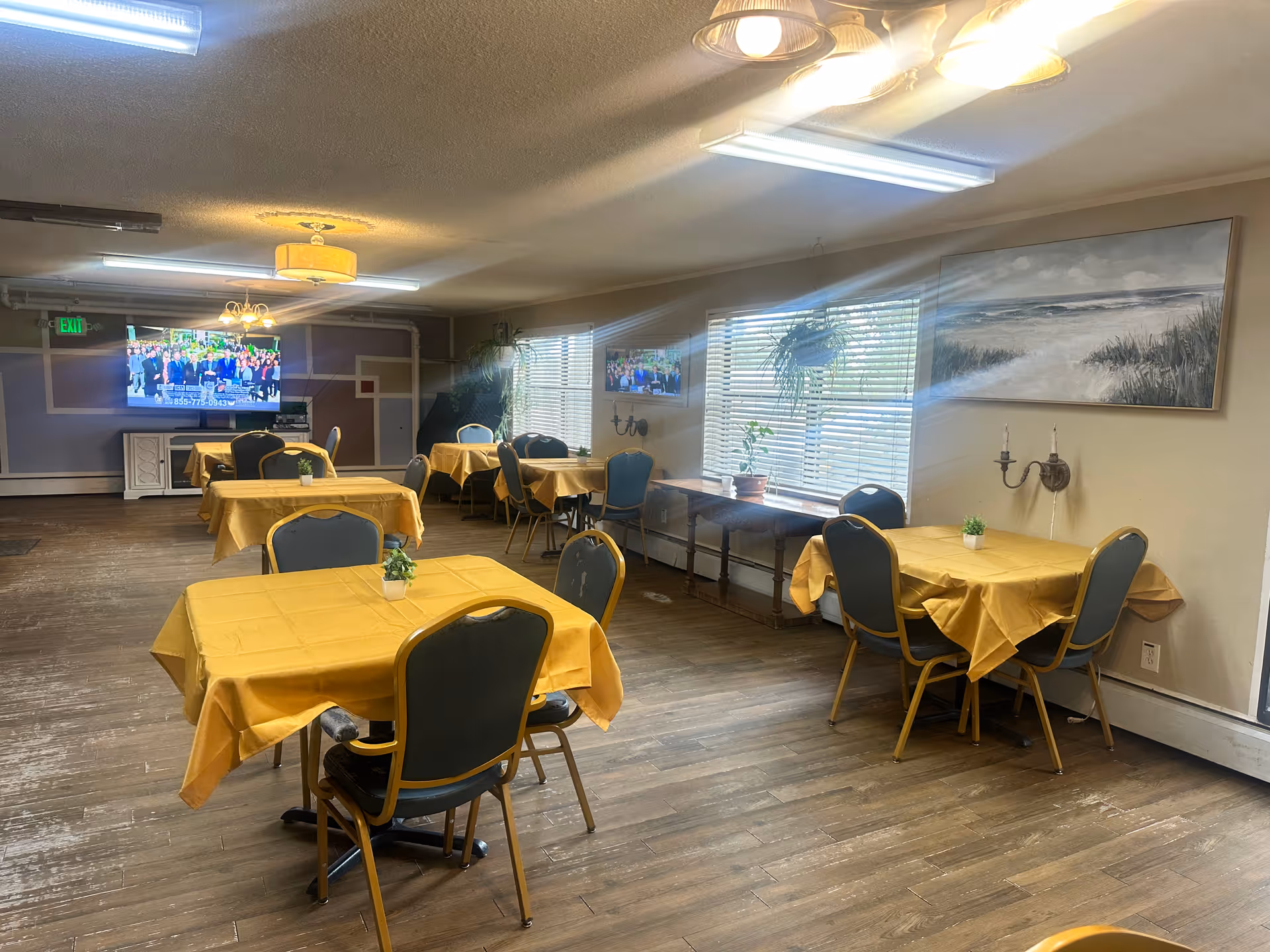 A dining room with several tables covered in yellow tablecloths, each surrounded by chairs. There are small potted plants on the tables. The room has wooden flooring, large windows with blinds, and a painting of a beach scene on the wall. A television is mounted on the far wall, and there are ceiling lights providing illumination.