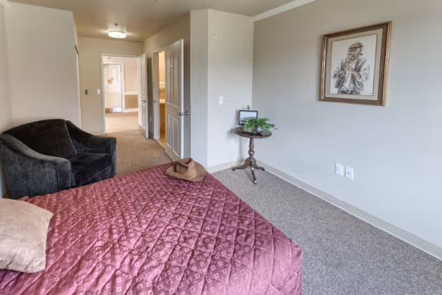 A senior living facility bedroom with a bed covered in a purple quilted bedspread, a beige pillow, and a brown hat on the bed. There is a dark upholstered armchair to the left, a small round wooden table with a plant and picture frame against the wall, and a framed artwork hanging above the table. The room has beige walls and carpeted floor, with an open door leading to a hallway.