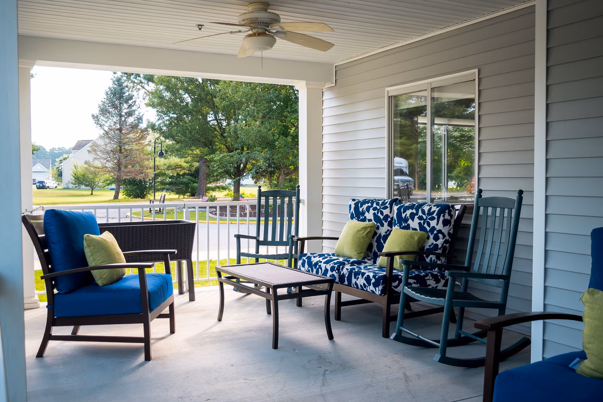 Covered outdoor patio area with cushioned seating including blue chairs with green pillows, a floral-patterned loveseat with green pillows, a small coffee table, and a ceiling fan. The patio overlooks a green lawn with trees and a street in the background.