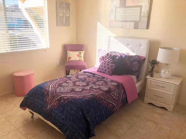Sunlit single bedroom with a patterned purple bedspread, white upholstered headboard, nightstand with lamp, and a chair by the window.