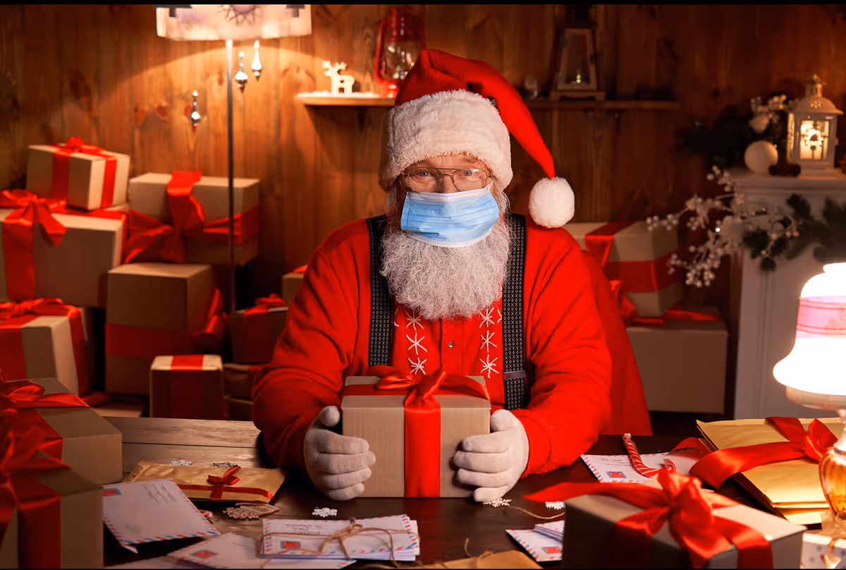 A person dressed as Santa Claus wearing a face mask and glasses, sitting at a table surrounded by wrapped Christmas presents with red ribbons. The setting is a cozy, warmly lit room with wooden walls and festive decorations.