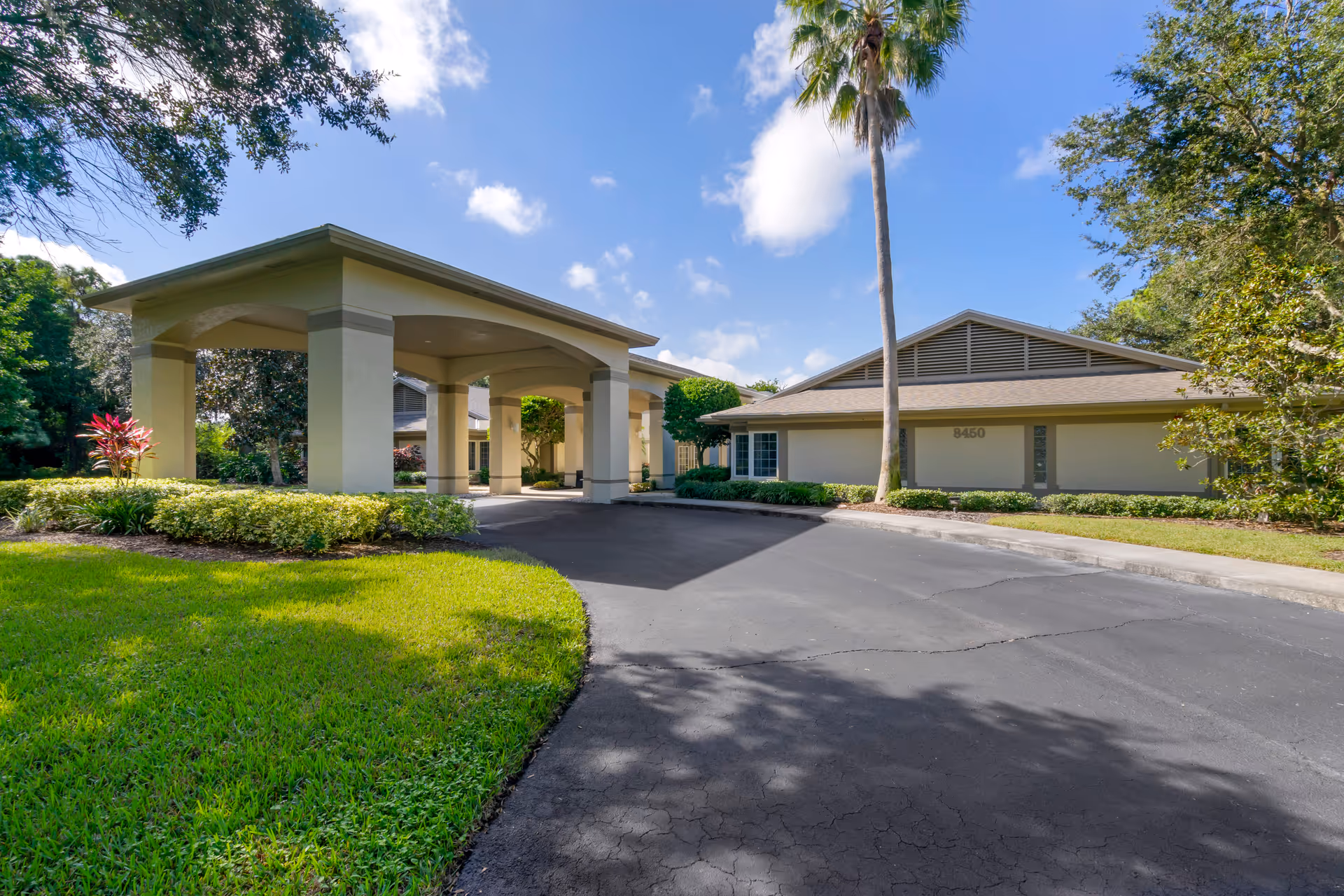 Entrance of a single-story senior living facility with a covered porte-cochere, driveway, palm tree, and landscaped lawn under a blue sky.