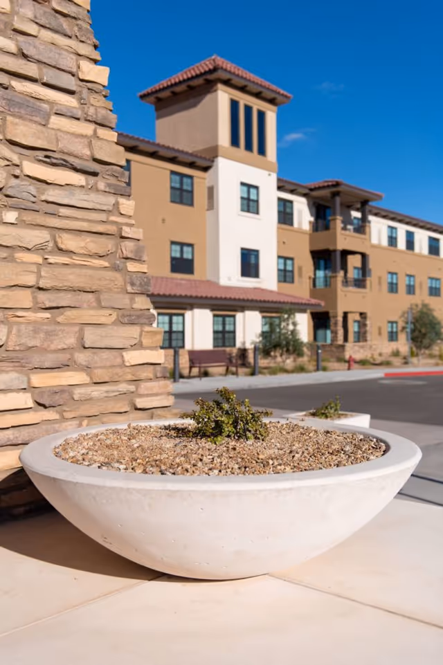A large round concrete planter filled with gravel and a small plant is in the foreground, with a stone wall to the left and a multi-story building with beige and white walls and red-tiled roofs in the background under a clear blue sky.