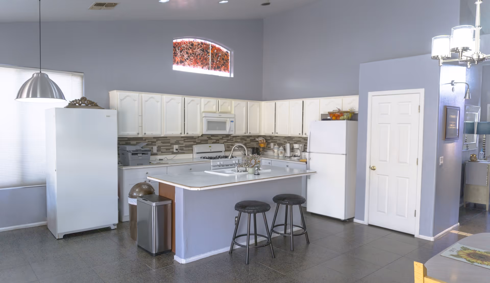 A bright kitchen with white cabinets, a white refrigerator, a white stove with a microwave above it, and a kitchen island with a sink and two black stools. The walls are painted light gray, and there is a small arched window above the cabinets. A silver pendant light hangs over the left side, and there are two trash bins near the island. The floor is tiled in gray.