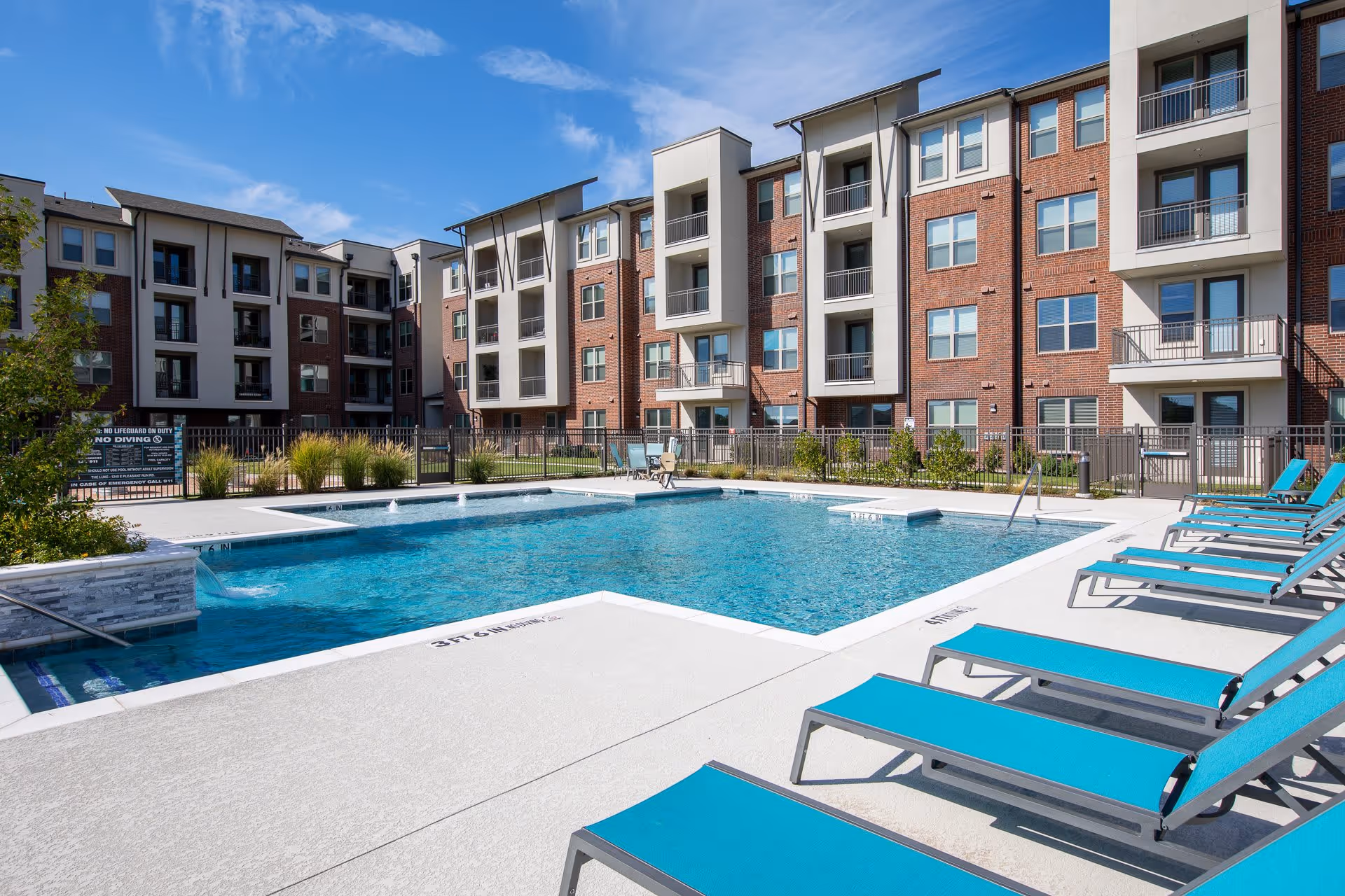 Outdoor swimming pool area with clear blue water surrounded by a concrete deck with multiple turquoise lounge chairs. In the background, there is a multi-story brick and beige apartment building with balconies and windows under a blue sky with some clouds.