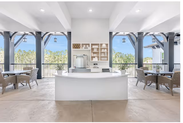 Outdoor covered patio area with a white central counter and shelving unit containing decorative items and a small refrigerator. There are multiple wicker chairs and tables arranged on either side of the counter, with black railings and large black support beams framing the open view of trees and blue sky in the background.