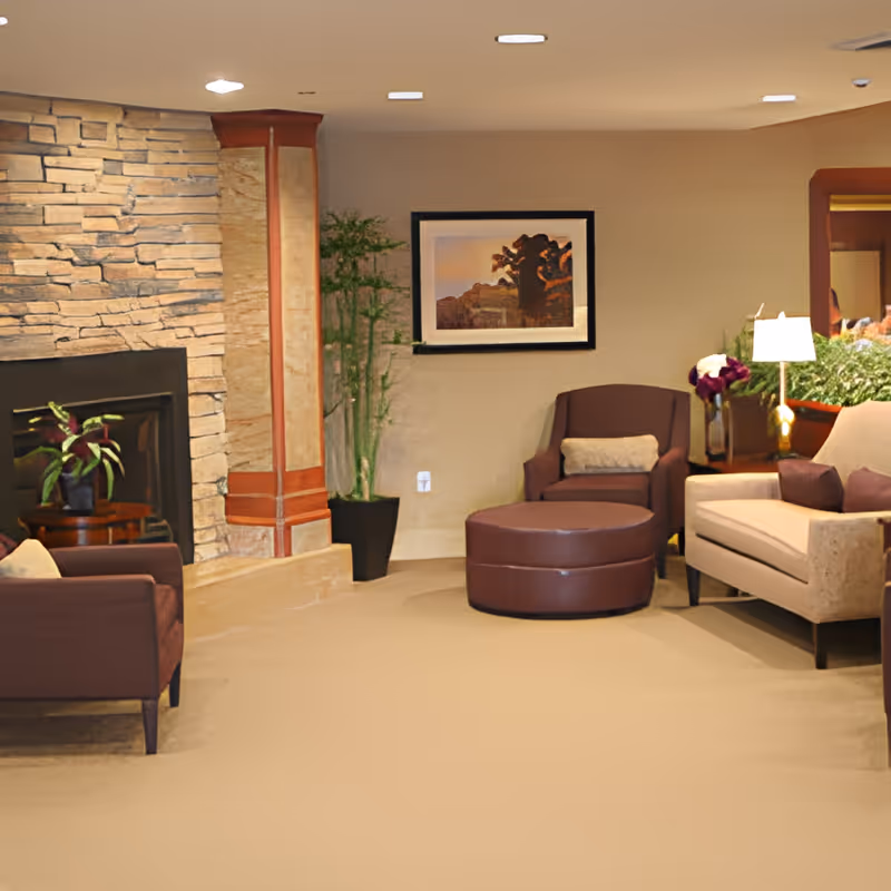 A cozy living room area with beige carpet, a stone fireplace on the left, a brown armchair with a matching ottoman, a beige sofa with purple cushions, a side table with a lamp and flowers, a potted plant, and a framed picture on the wall.