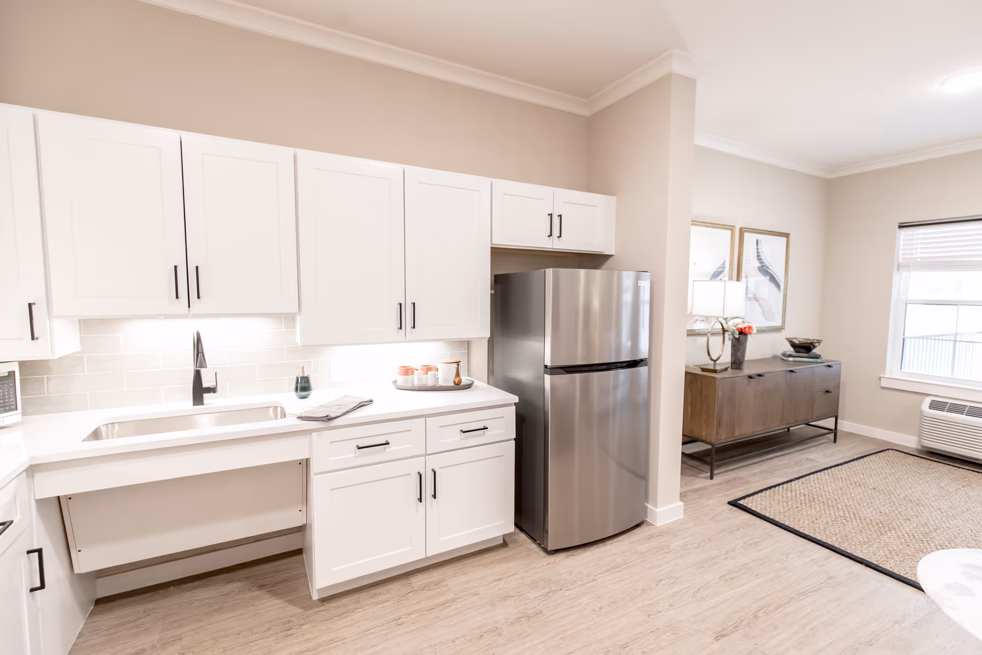 Bright kitchen area with white cabinets, stainless fridge, sink, and adjacent living space.