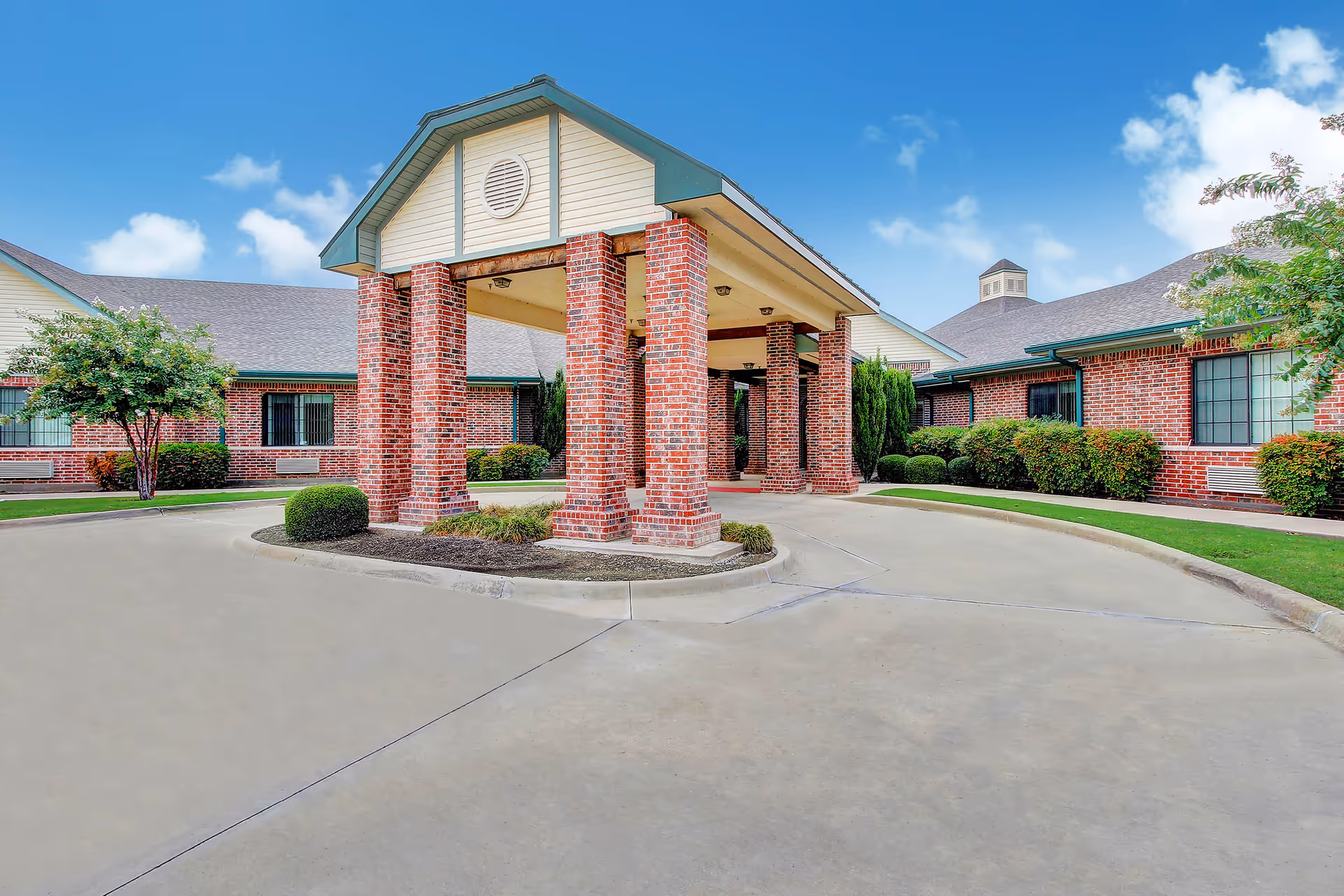 Exterior view of a senior living facility entrance with a covered drop-off area supported by brick columns, surrounded by well-maintained landscaping and a clear blue sky.