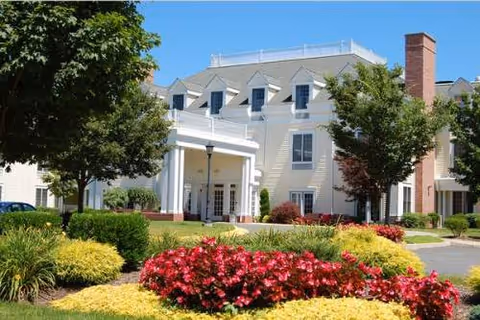 White multi-story senior residence with entry canopy, brick chimney, and colorful landscaped front yard.