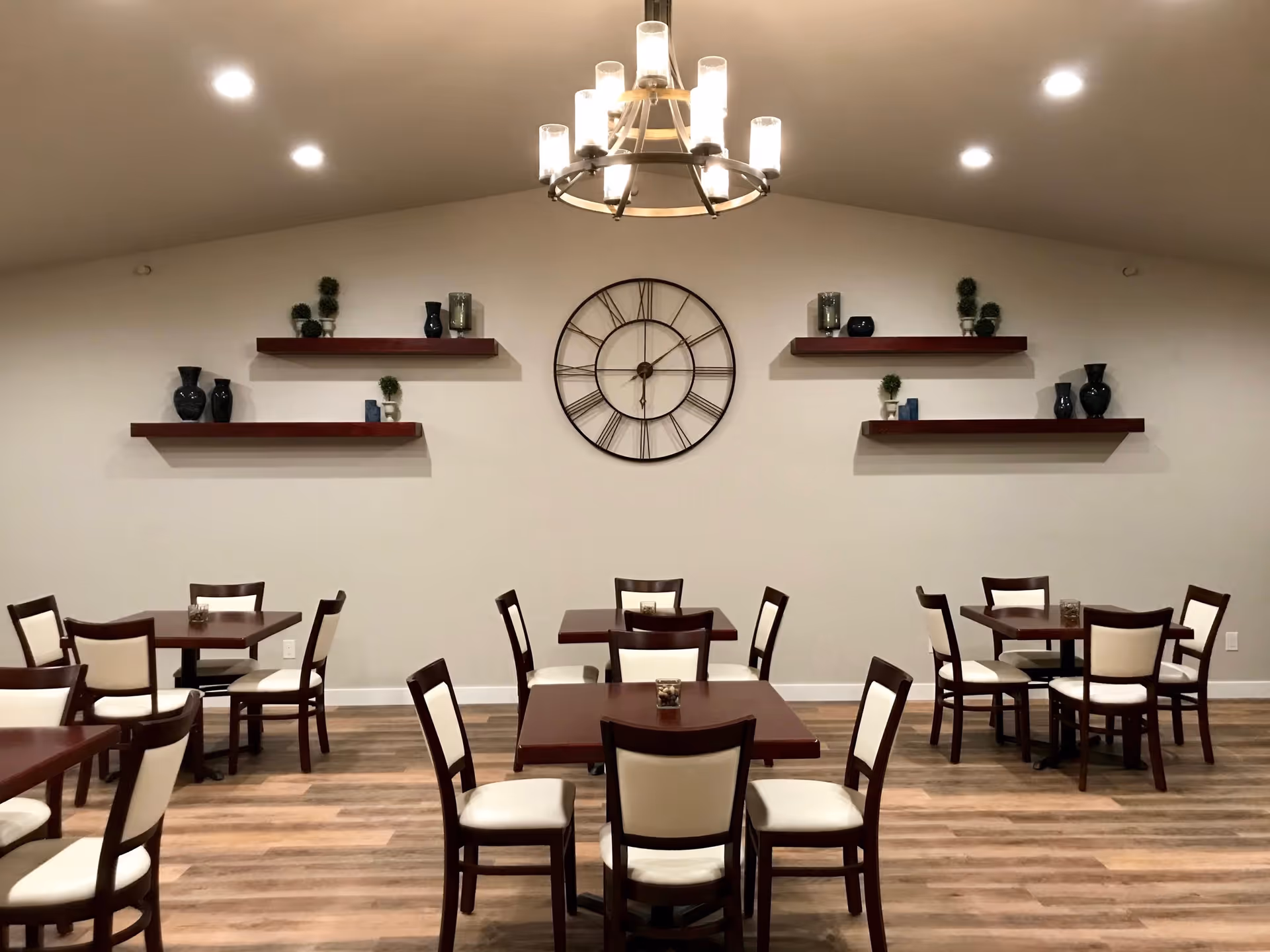 A dining room with several wooden tables and chairs with white cushions. The walls are light-colored with four wooden shelves decorated with small plants and vases. A large round clock with Roman numerals is centered on the wall. A chandelier with multiple lights hangs from the ceiling, and recessed lighting is also visible.
