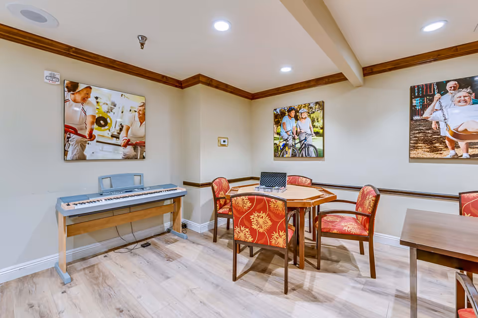 A room with a wooden floor and beige walls featuring three framed photos of seniors engaging in activities. There is a digital keyboard against one wall and a wooden table with four red and yellow floral-patterned chairs around it. The room is well-lit with recessed ceiling lights.