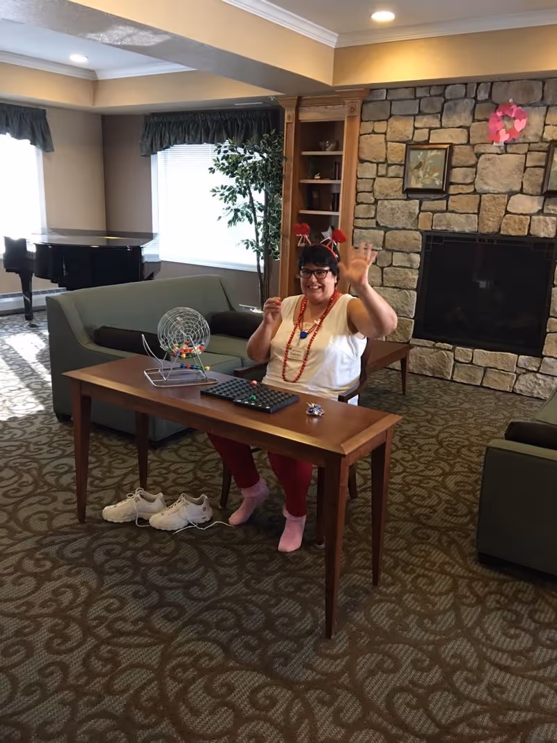 A woman wearing red pants, a white shirt, and festive red accessories is sitting at a wooden table in a cozy living room. She is smiling and waving at the camera. On the table are a bingo cage with balls and a bingo board. Behind her is a stone fireplace with framed artwork and a pink heart decoration. The room has patterned carpet, green sofas, a bookshelf, a potted plant, and a grand piano near the windows with green valances.