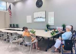 A bright community dining/meeting room with long tables, floral centerpieces, an American flag, wall clock and artwork, and several seated elderly residents.