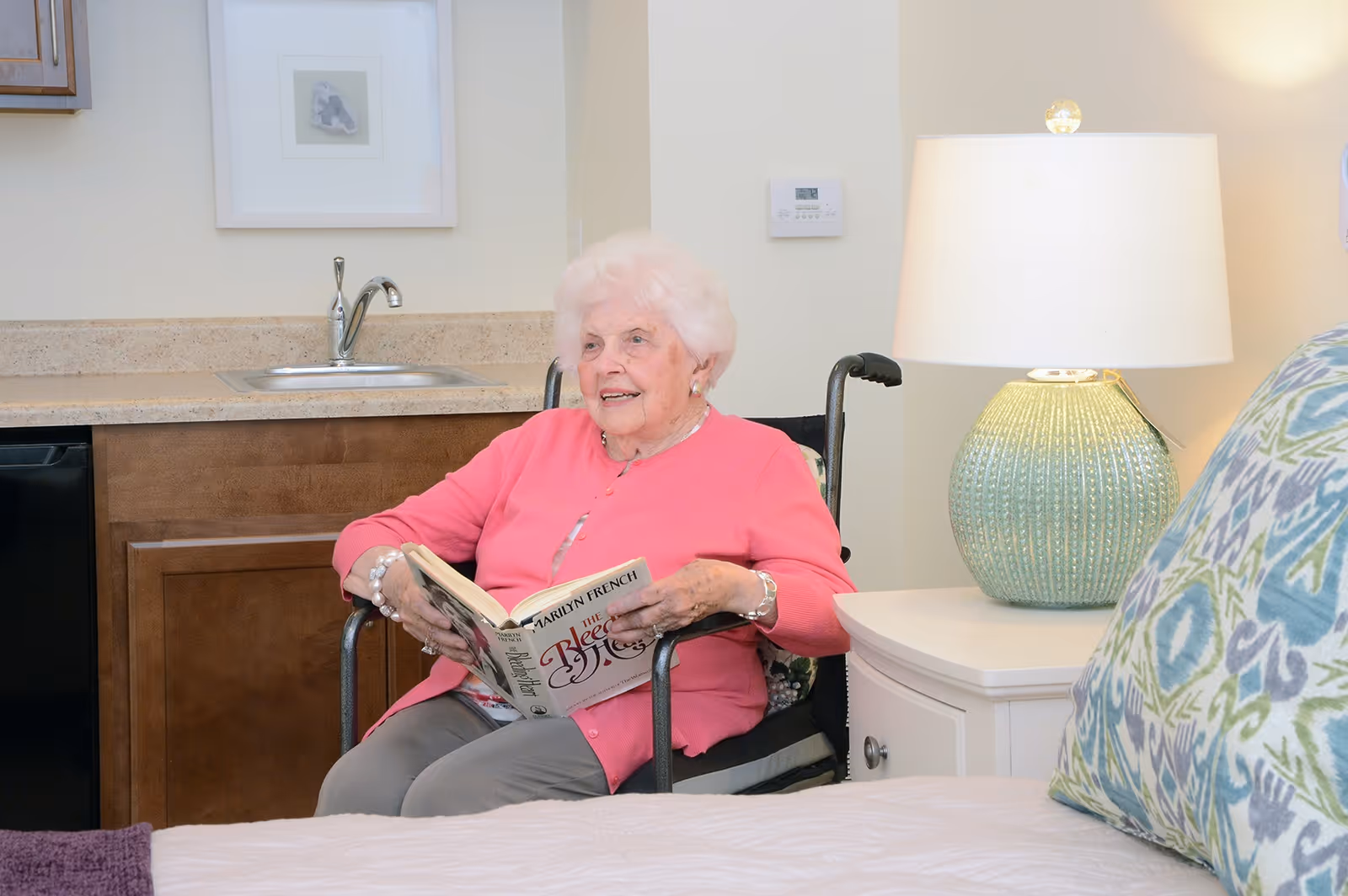 An elderly woman with white hair wearing a pink cardigan sits in a wheelchair in a cozy room. She is holding and reading a book titled 'The Bleeding Heart' by Marilyn French. Behind her is a small kitchenette with a sink and cabinets. To her right is a white nightstand with a green textured lamp and a bed with a colorful patterned pillow in the foreground.