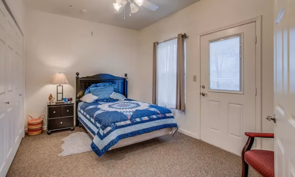 A bedroom with a single bed covered in a blue patterned quilt, two pillows, a black headboard, a nightstand with a lamp and small decorative items, a window with curtains, a door with a window, a red chair, and a carpeted floor.