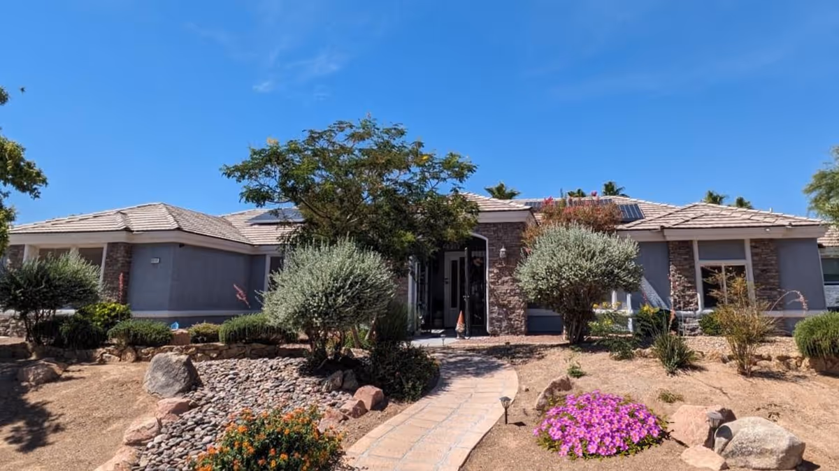 Single-story ranch-style senior home with stone accents and solar panels, a paved walkway leading through drought-tolerant landscaping under a clear blue sky.