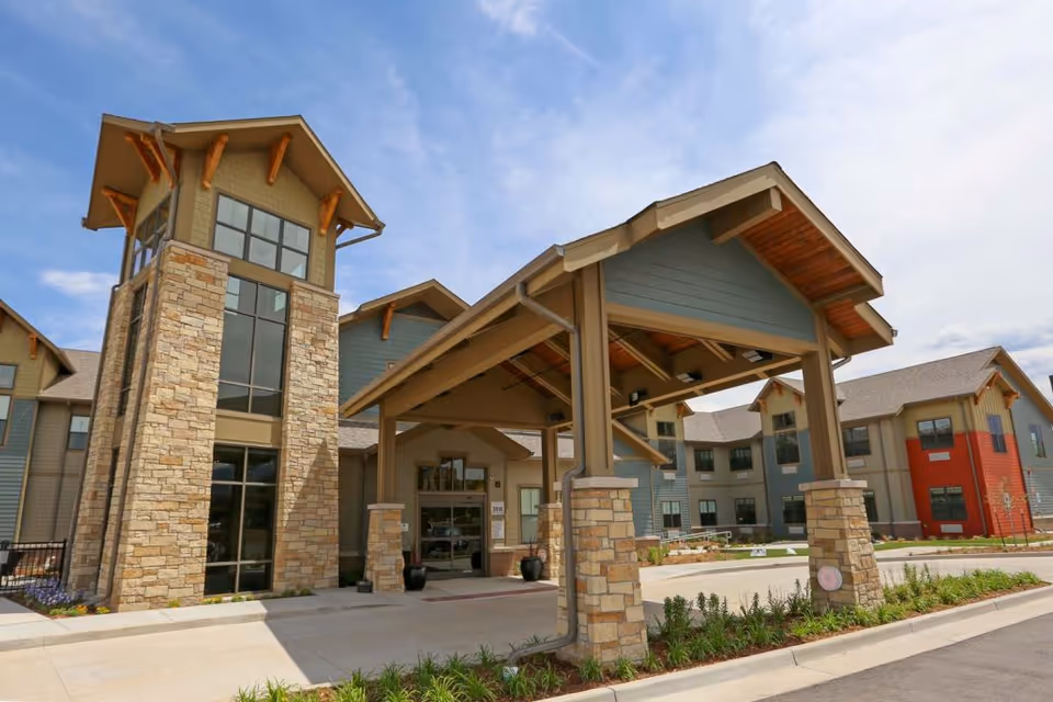 Exterior view of The Healthcare Resort Of Colorado Springs building featuring a covered entrance supported by stone pillars, large windows, and a mix of stone and siding on the facade under a partly cloudy sky.
