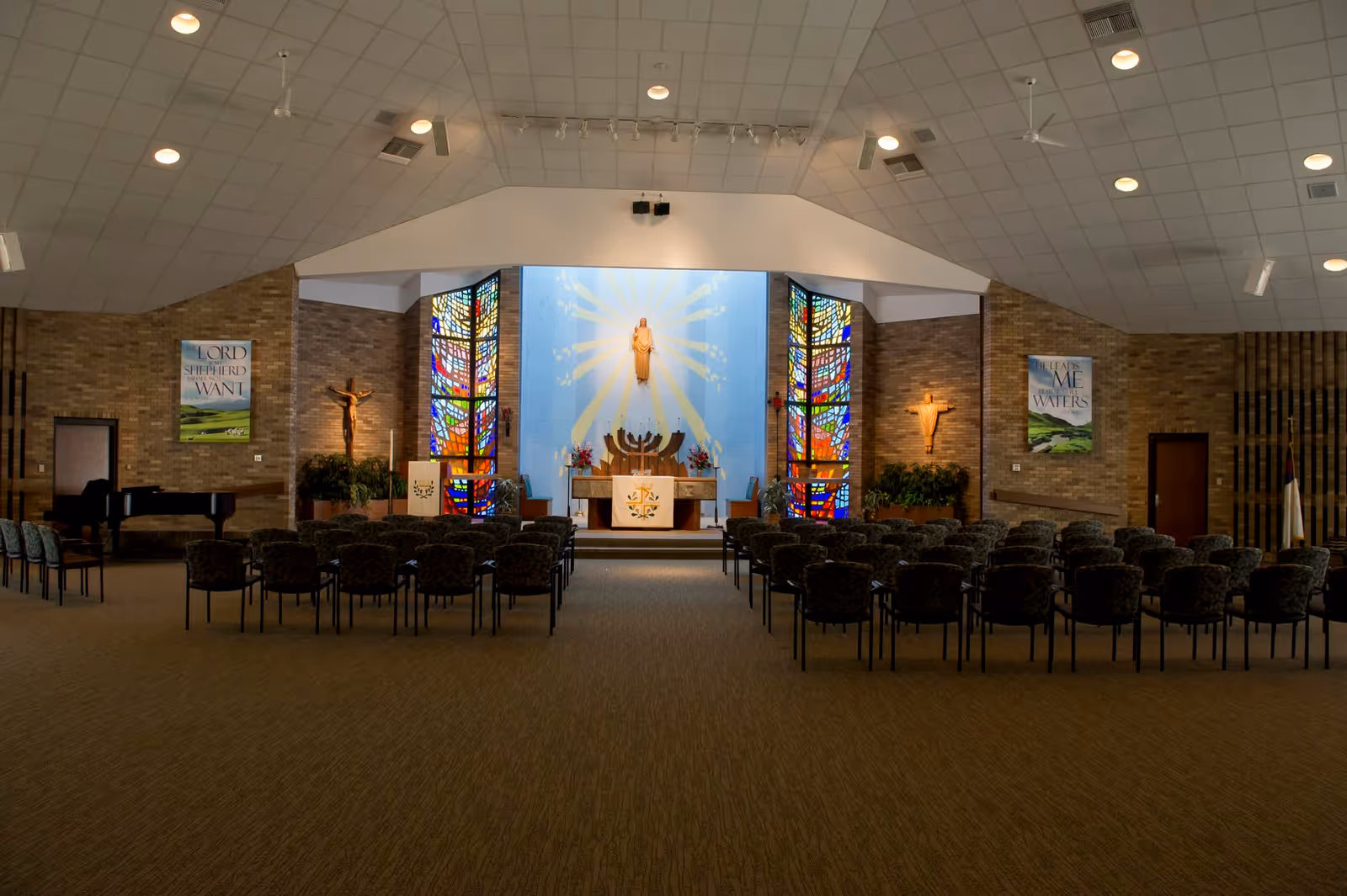 Interior view of a chapel or worship room with rows of chairs facing an altar. The altar area features stained glass windows on either side, a statue of Jesus on the wall with rays of light painted behind it, and religious symbols including a menorah and crucifixes. There are two posters on the brick walls with religious quotes, a piano on the left side, and ceiling lights illuminating the space.